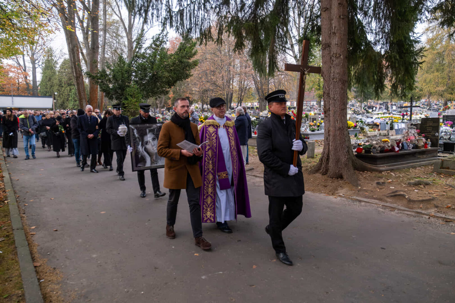 Kondukt pogrzebowy z księdzem i krzyżem w alei cmentarza